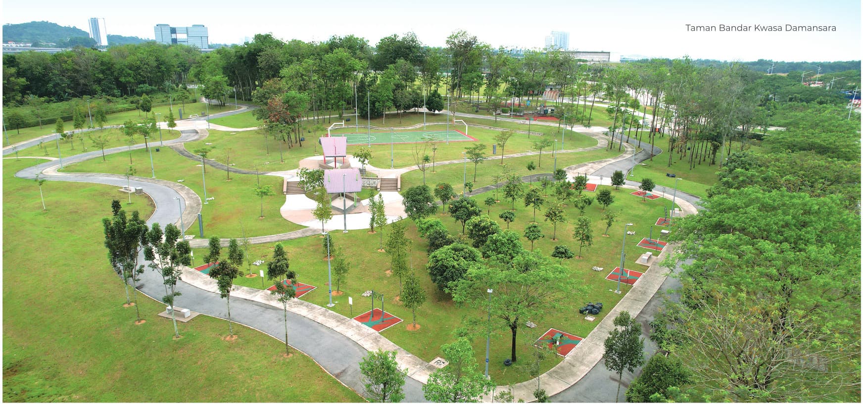 Taman Bandar Kwasa Damansara from above — 42 acres of lawn, young trees, looping footpaths, a playground and a covered court, with the Kwasa Damansara skyline beyond