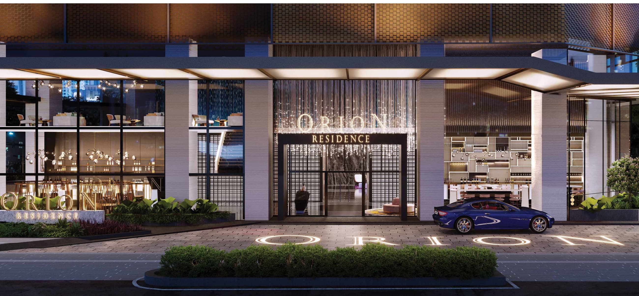 Orion Residence ground-floor arrival — the lit Orion Residence portal between retail frontages, with the drop-off, landscaped verge and the projected Orion lettering on the driveway