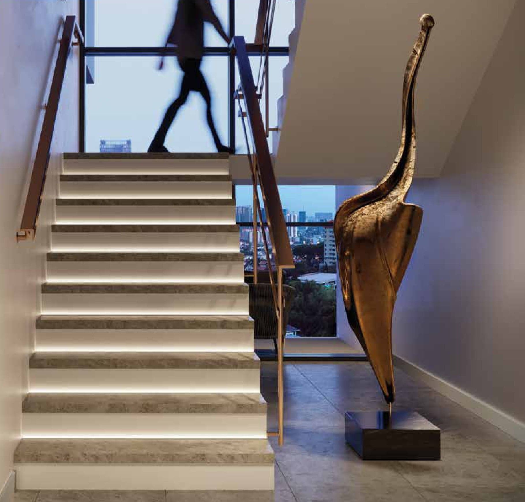 The internal staircase in a duplex, lit under every tread, with the city through the landing window
