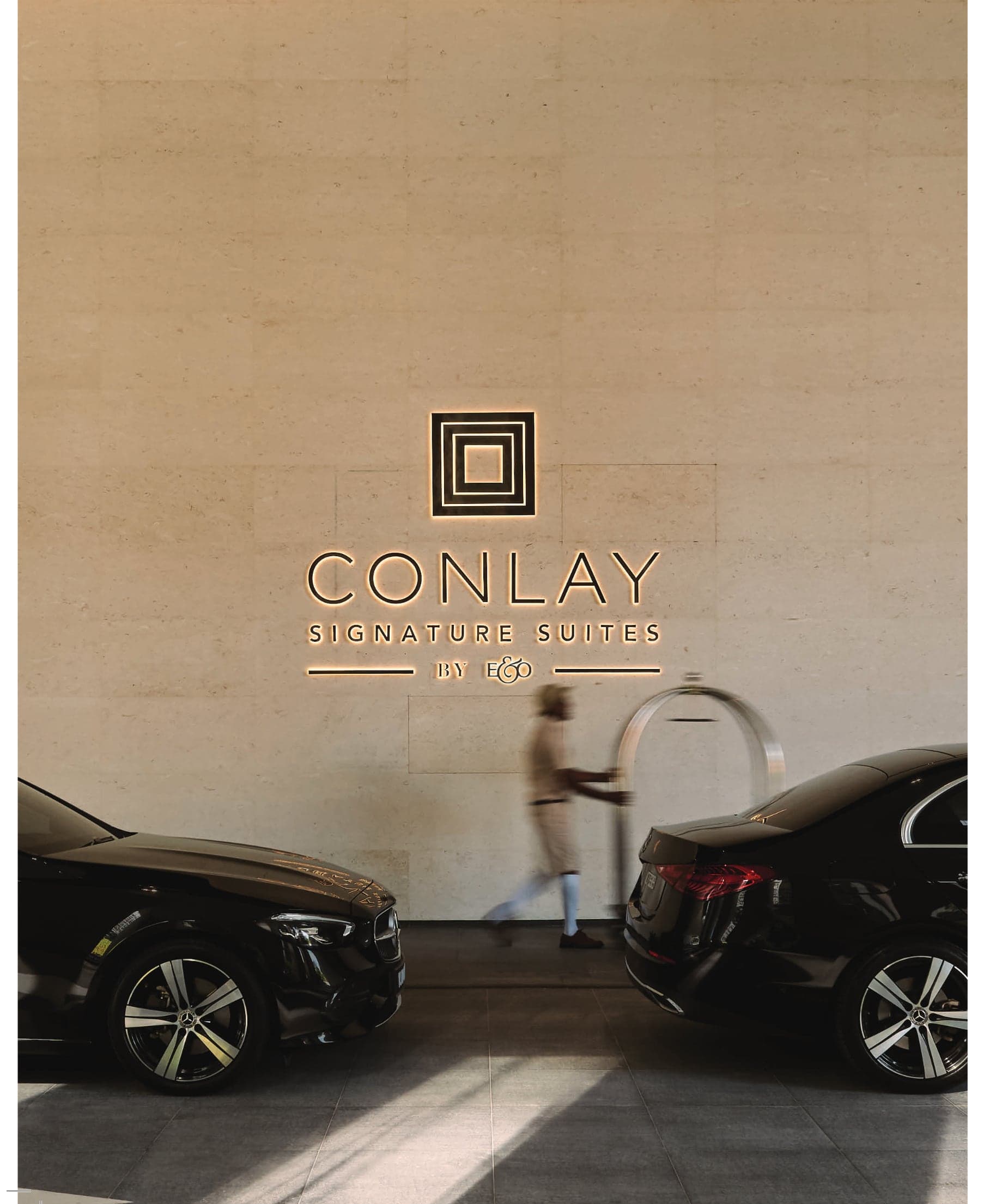 The Conlay arrival court — the Conlay Signature Suites by E&O brass lockup on the stone porte-cochère wall, with cars drawn up and a porter passing between them